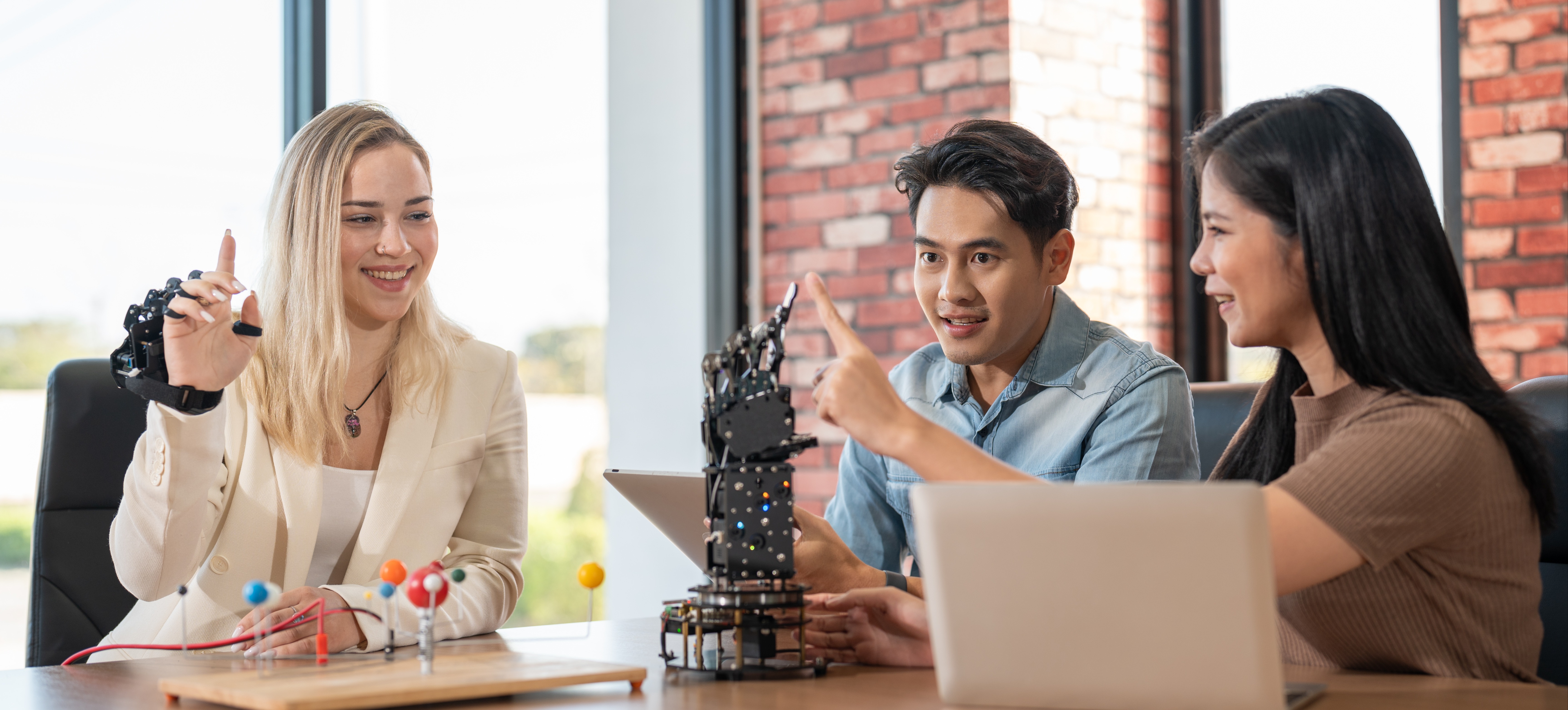[Feature Image] Three college learners sit around a wood-topped table testing their latest project, which is part of their class on how to learn robotics.
