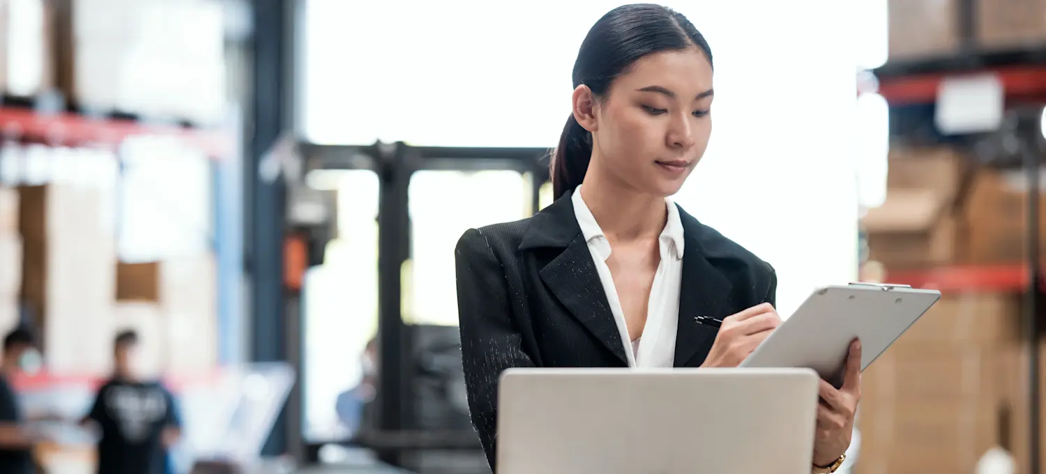 [Featured Image] An inventory control analyst looks at a laptop while holding a clipboard in a warehouse.
