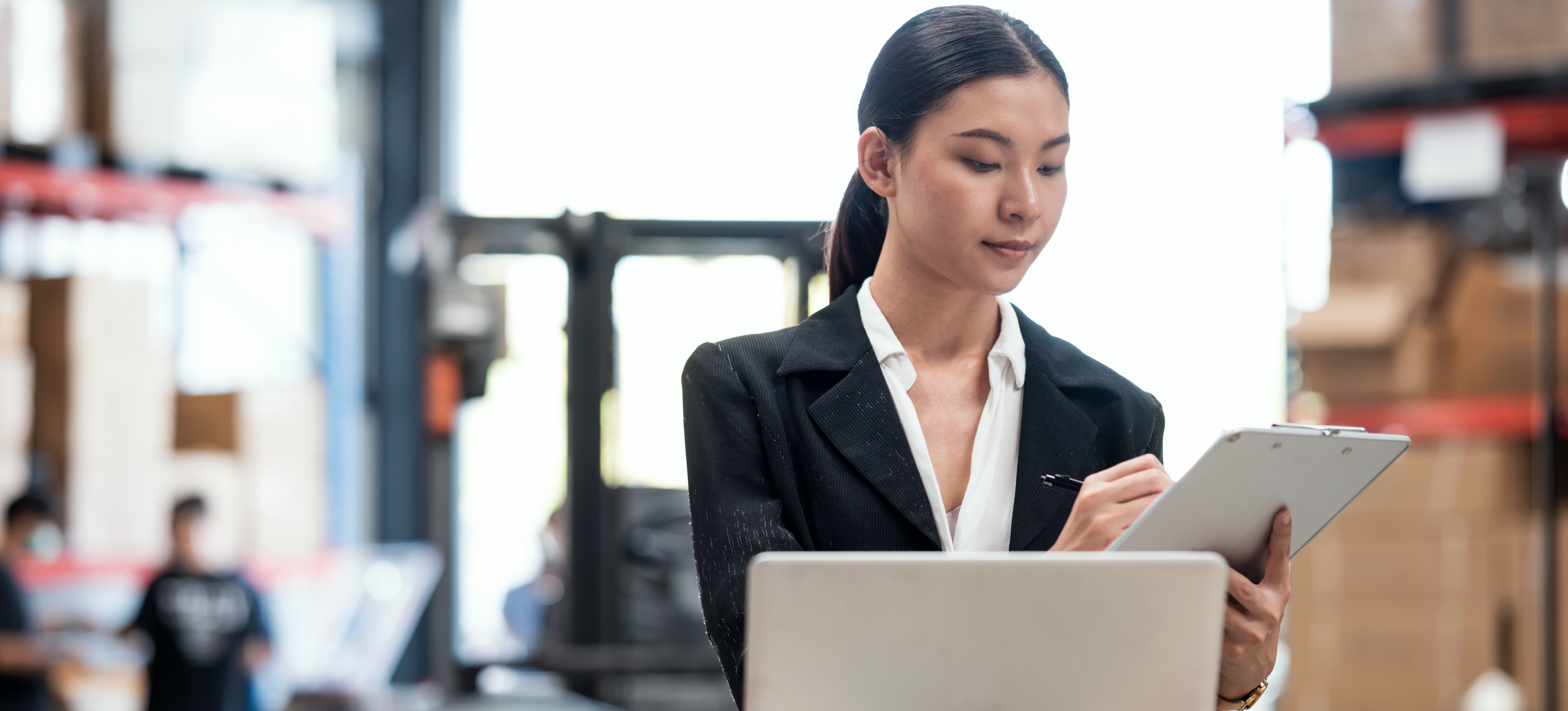 [Featured Image] A woman inventory control analyst looks at a laptop while holding a clipboard in a warehouse.
