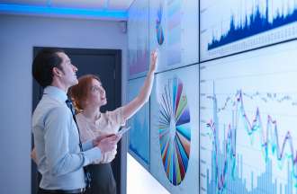 [Featured Image] Two people examine data represented in graphs and charts on large screens in an office.