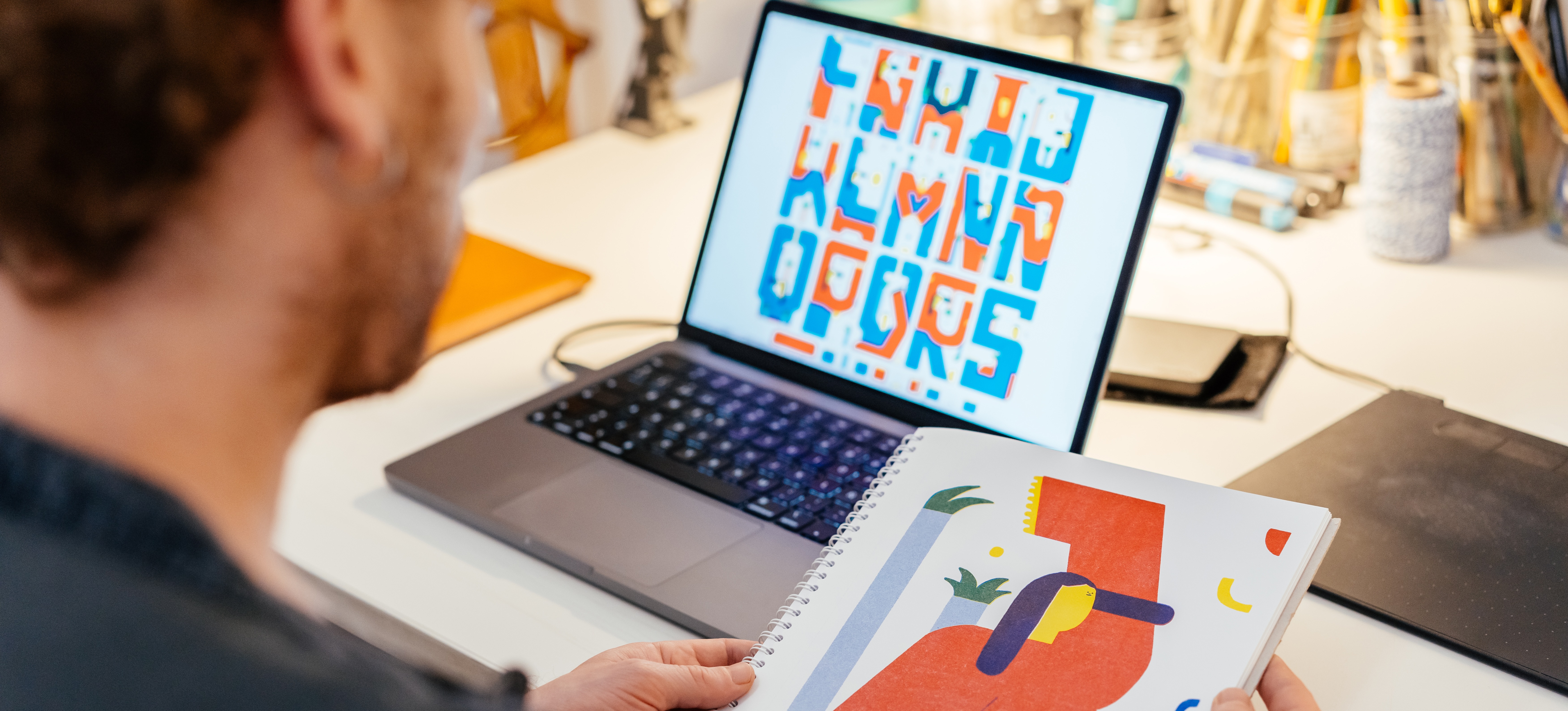 [Featured Image] An artist views a hand-drawn image in an art journal while experimental typography is displayed on their laptop screen.
