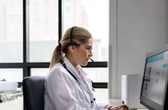 [Featured Image]:  A female telehealth nurse, wearing a white uniform, is sitting in front of her computer, communicating with a patient.