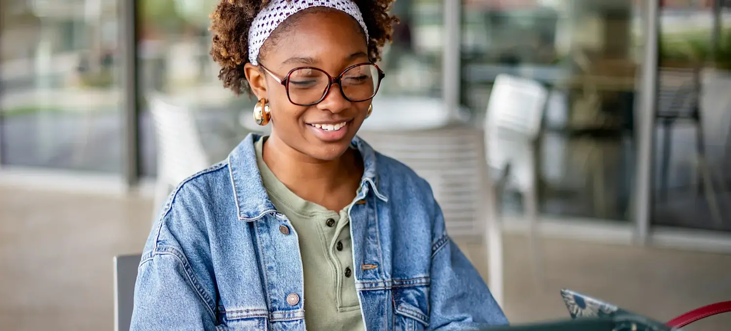 [Featured Image] A smiling student is using ChatGPT for studying on their laptop while sitting in an outdoor public patio area.
