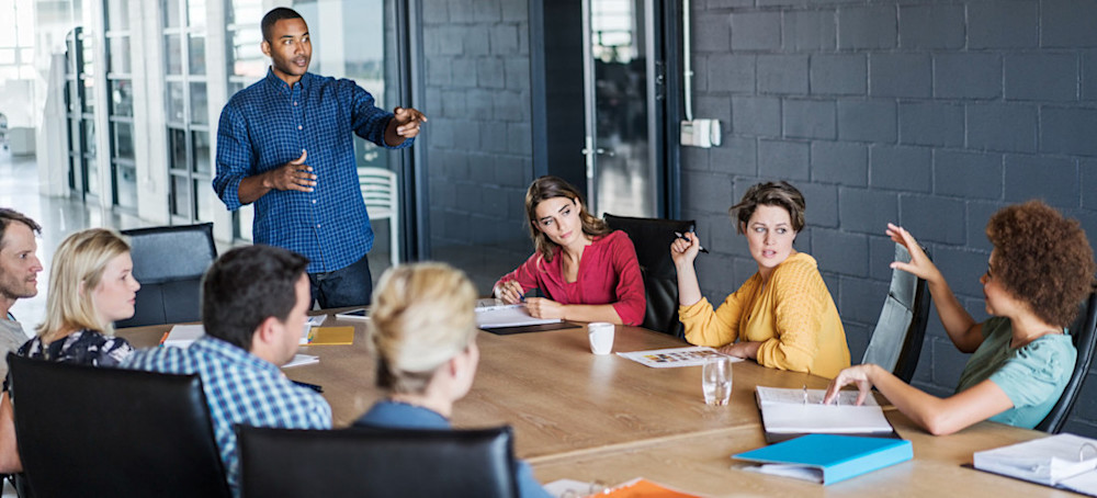 [Featured Image] A businessman stands at a table and points to one of his colleagues seated around the table.