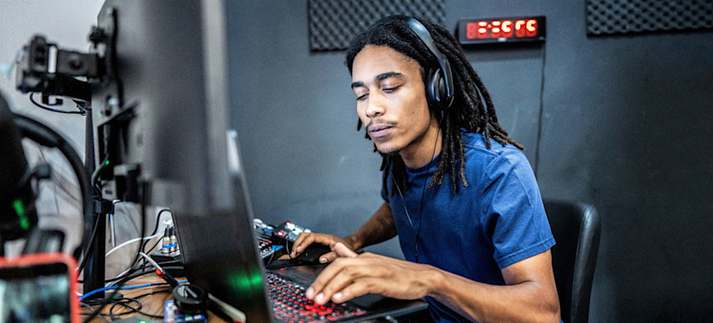 [Featured Image] A technician wearing headphones works on audio engineering as they sit in front of their computer and equipment in their sound studio.
