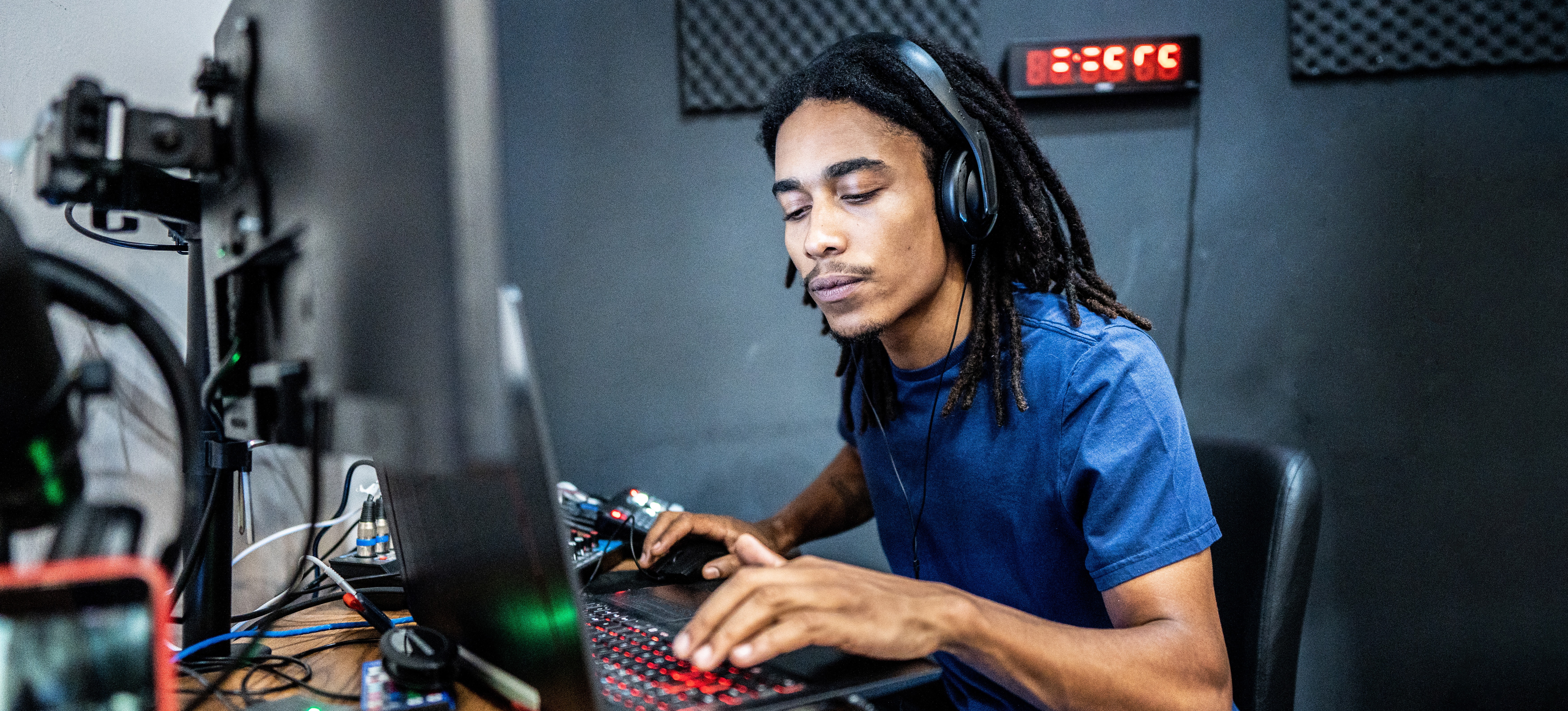 [Featured Image] A technician wearing headphones works on audio engineering as they sit in front of their computer and equipment in their sound studio.
