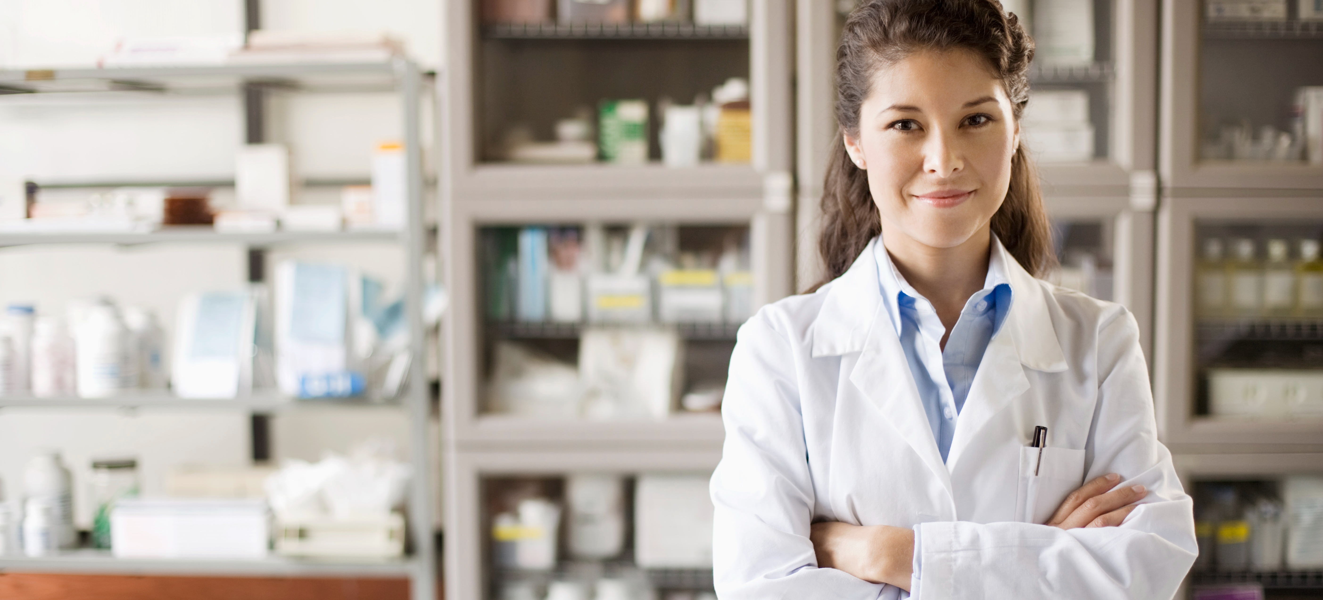 [Featured image] A smiling pharmacist stands behind a counter with her arms crossed with shelves of medication behind her.