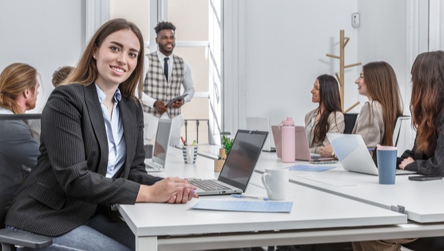 [Featured image] A data analyst sits at their laptop smiling at the camera with a group of coworkers in the background.