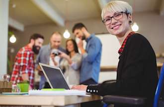 [Featured image] A data analyst sits at their laptop smiling at the camera with a group of coworkers in the background.