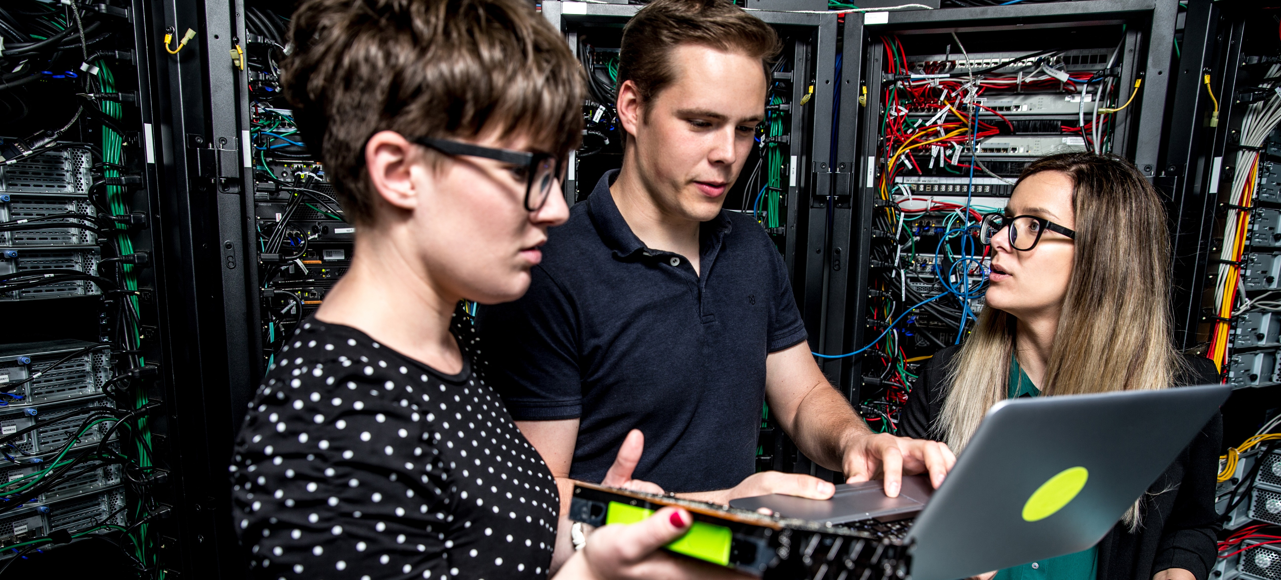 [Featured Image] Three data center technicians stand in a crowded server room amid much equipment and wiring as one scans data on a laptop.
