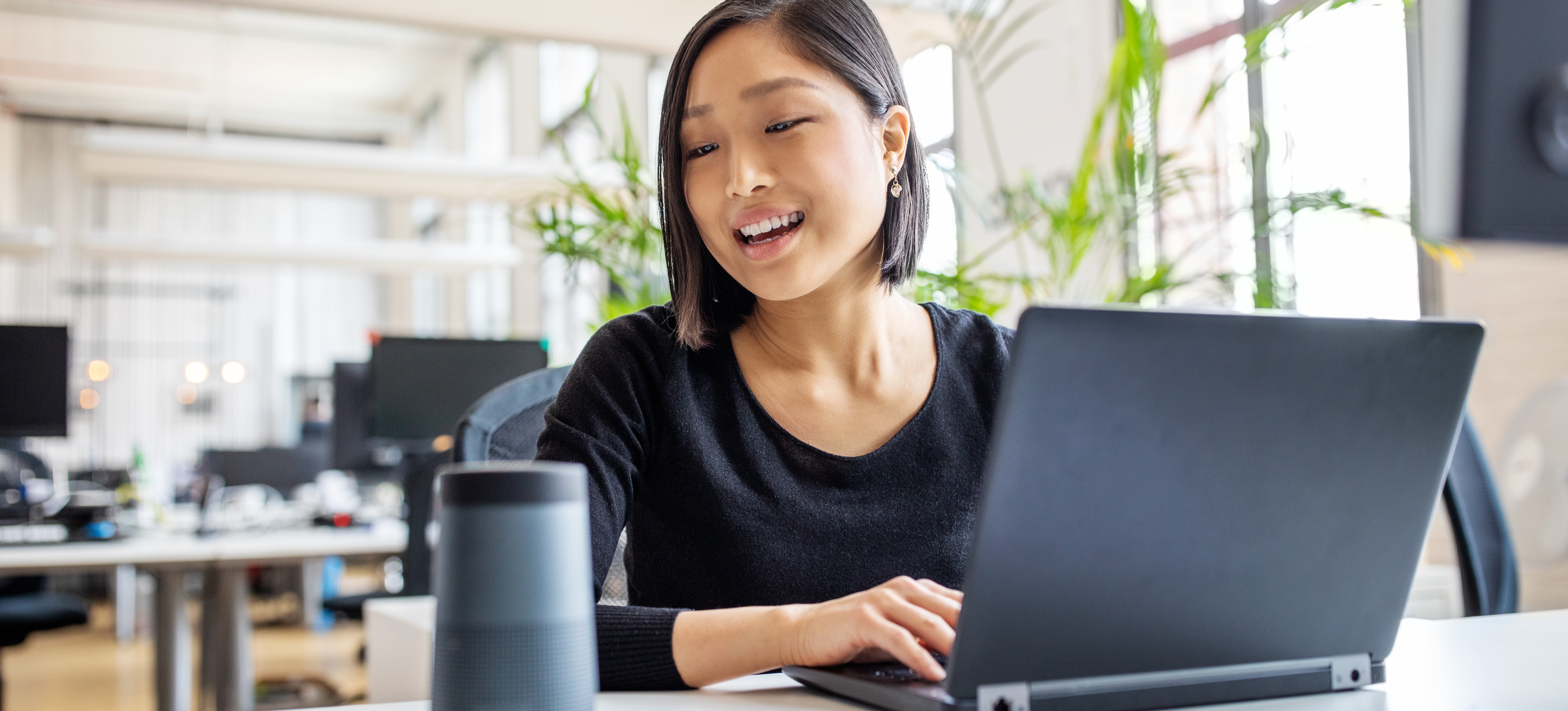 [Featured Image] A businesswoman types on her laptop while speaking into a voice assistant device, which uses a type of artificial neural network called natural language processing.
