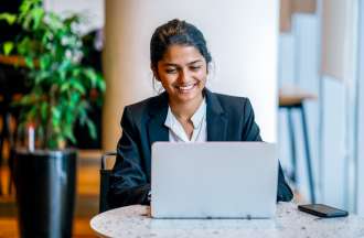 [Featured image] Smiling woman in a business suit sitting at a table with her laptop and smartphone.