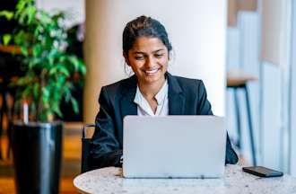 [Featured image] Smiling woman in a business suit sitting at a table with her laptop and smartphone.