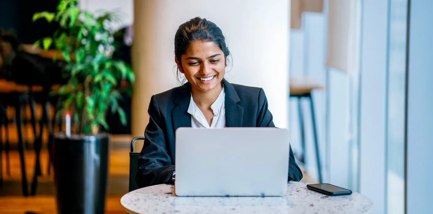 [Featured image] Smiling person in a business suit sitting at a table with their laptop and smartphone.