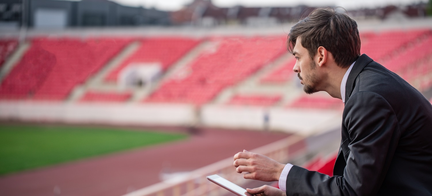 [Featured Image] A sports manager looks over a soccer field from the bleachers to brainstorm ideas for the team's upcoming roster
