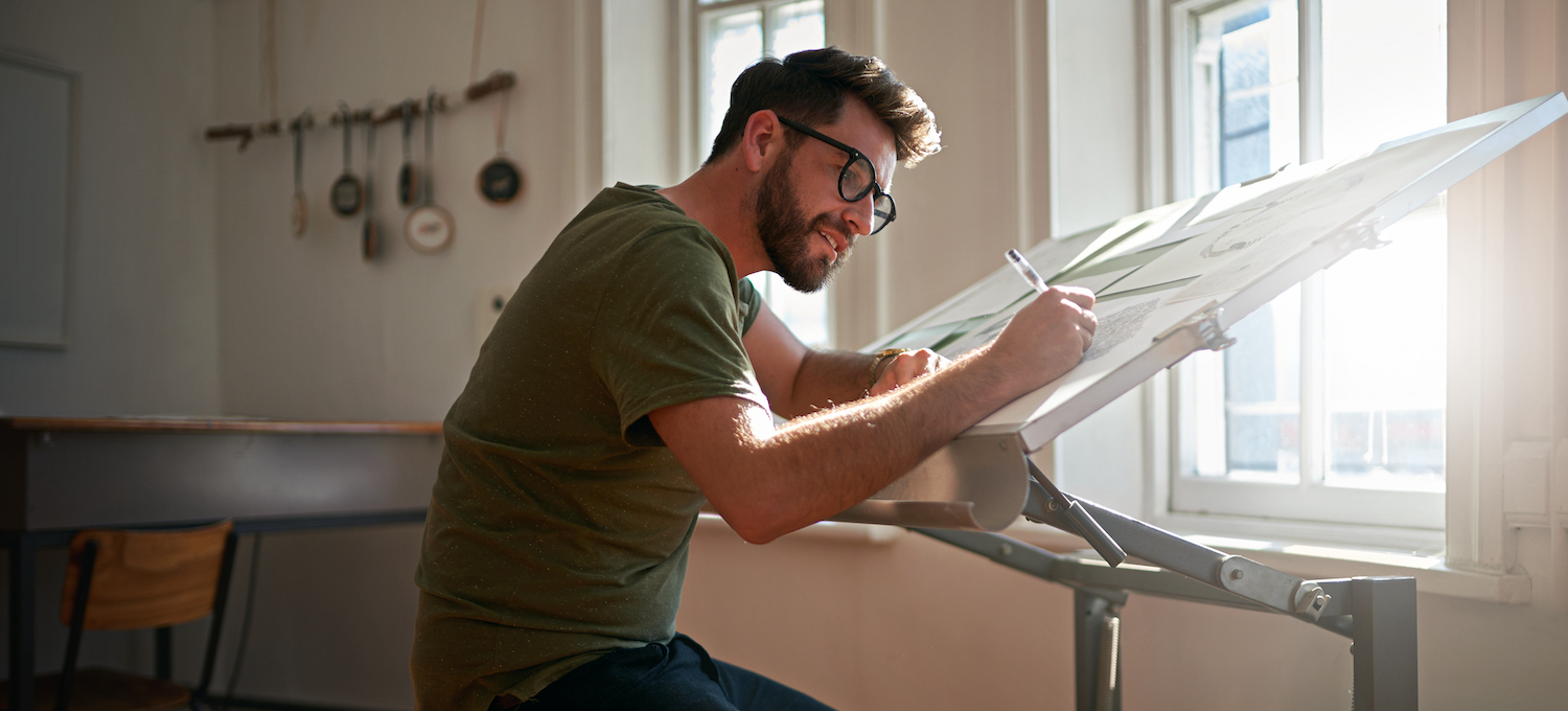 [Featured image] A graphic design student sits at a tilted table and works on a new design project. They have a series of papers in front of them and are using a pen to update their designs.