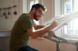 [Featured image] Graphic design student sits at a tilted table and works on a new design project. He has a series of papers in front of him and is using a pen to update his designs.