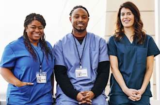 Three nurses, two female and one male all wearing blue scrubs, smile in a hospital.