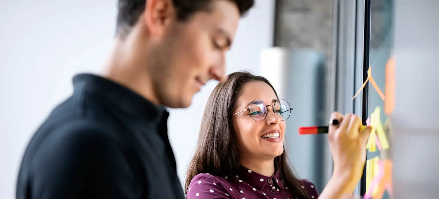 [Featured Image] Two people are placing post-it notes on a whiteboard. 