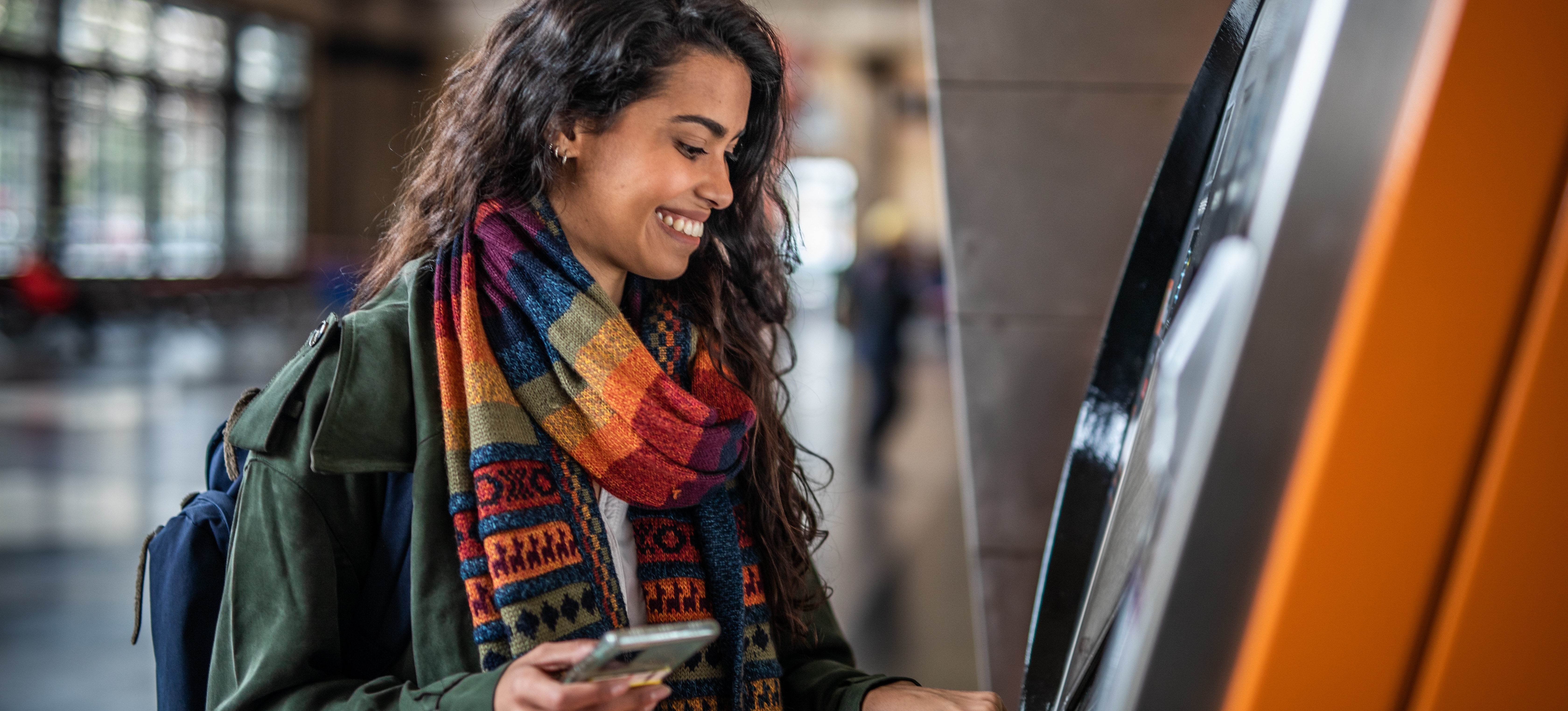 [Featured Image] A smiling person with long brown hair wearing a colorful scarf holds their smartphone and takes money from an ATM as part of their annual income.
