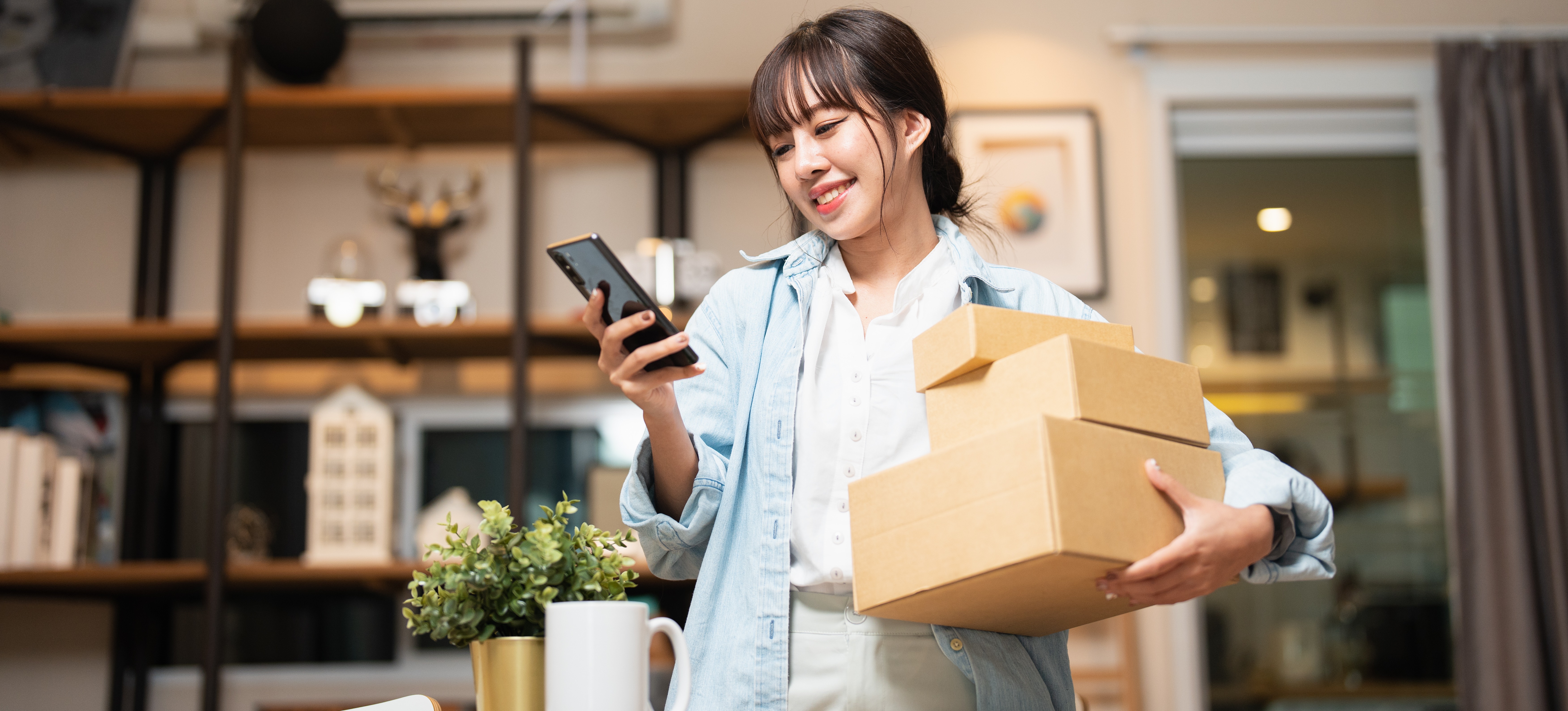 [Featured image] A customer holds three brown packages in their arm while looking at their phone. They're standing in a living room.