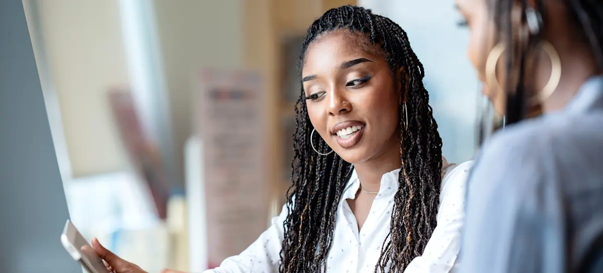 [Featured Image] Two people sitting side by side at a desk, while looking at plans on a tablet.
