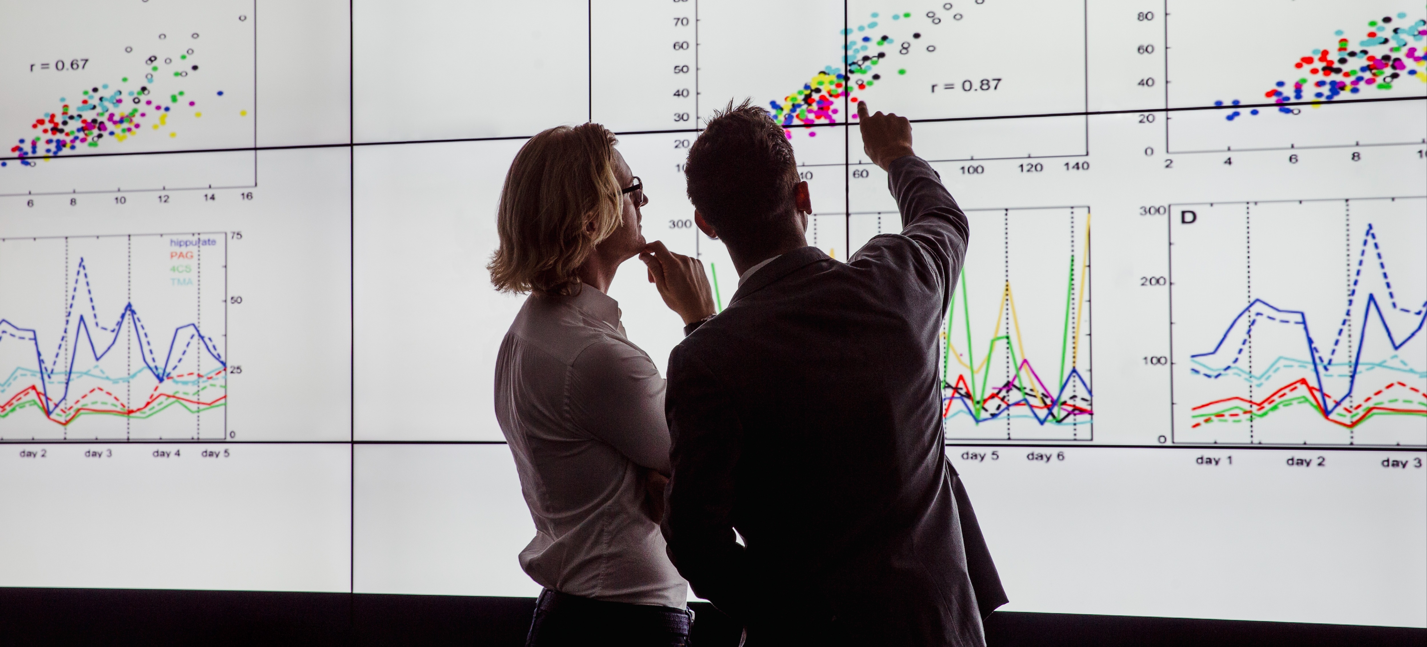[Featured Image] In a dark room, one man explains ETL to another while they examine a data visualization. 
