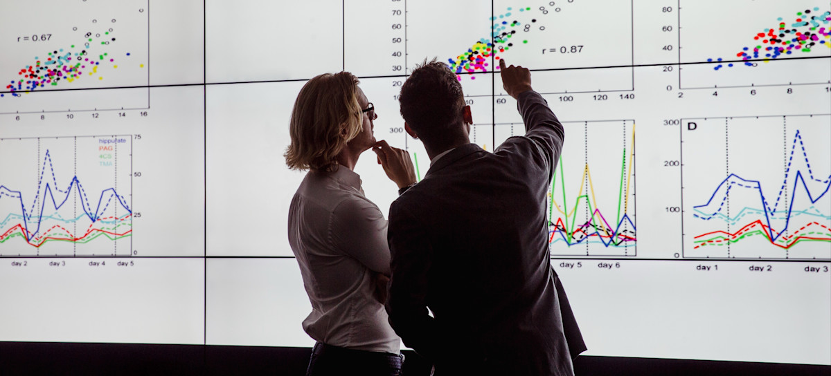 [Featured Image] In a dark room, one man explains ETL to another while they examine a data visualization. 
