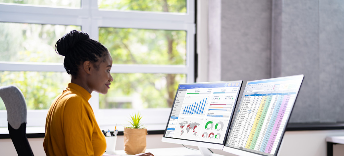 [Featured Image]: A person wearing an orange dress is sitting in front of two computer screens as they work as an entry-level data analyst. 