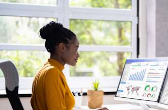 [Featured Image]: A woman wearing an orange dress, is sitting in front of two computer screens as works as an entry-level data analyst. 