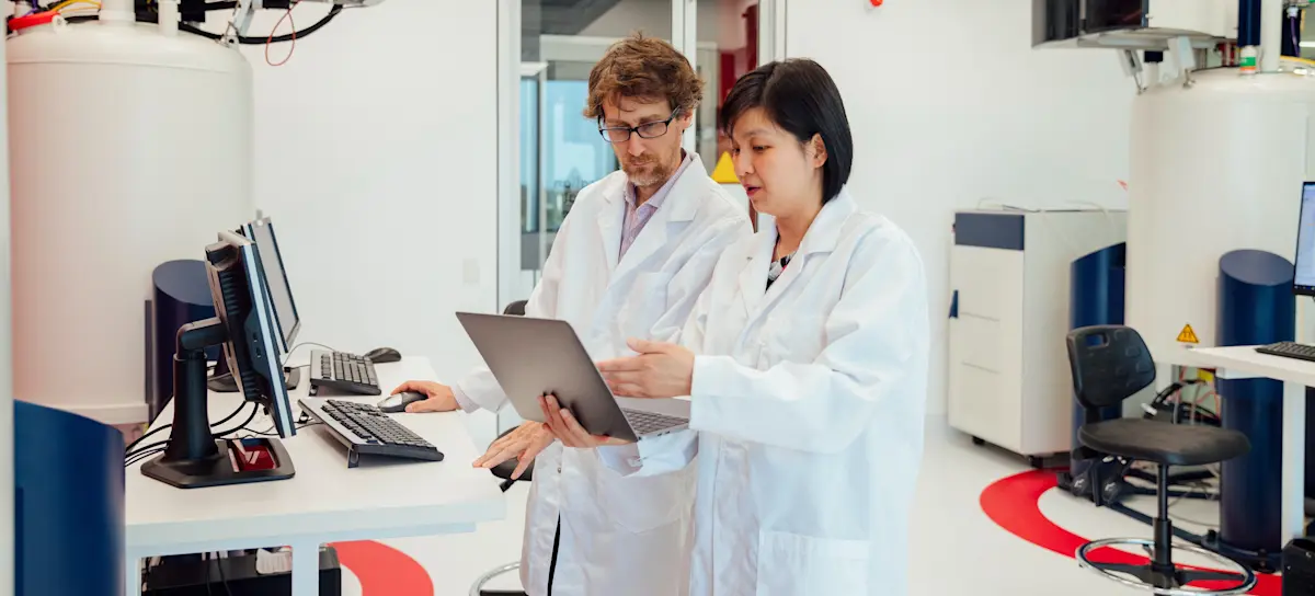 [Featured image] A physician assistant in a white lab coat shows test results on a laptop to their colleague.