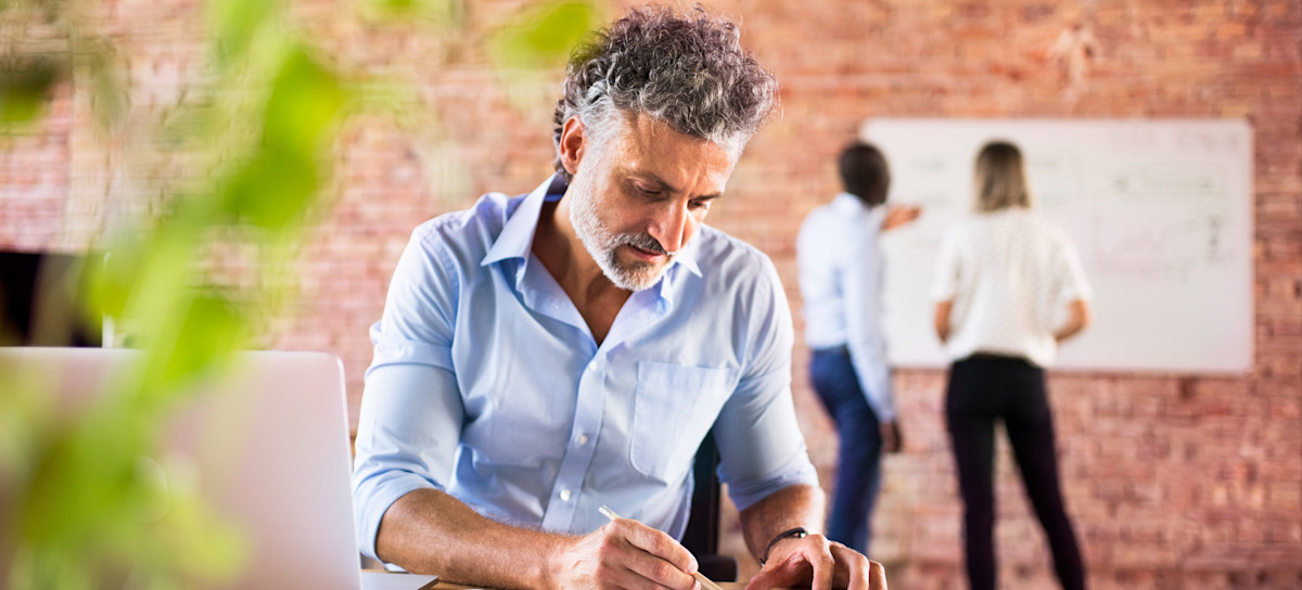 [Featured Image] A project manager reads earned value management results as they sit at a table with a laptop and designs, with two team members looking at designs in the background.
