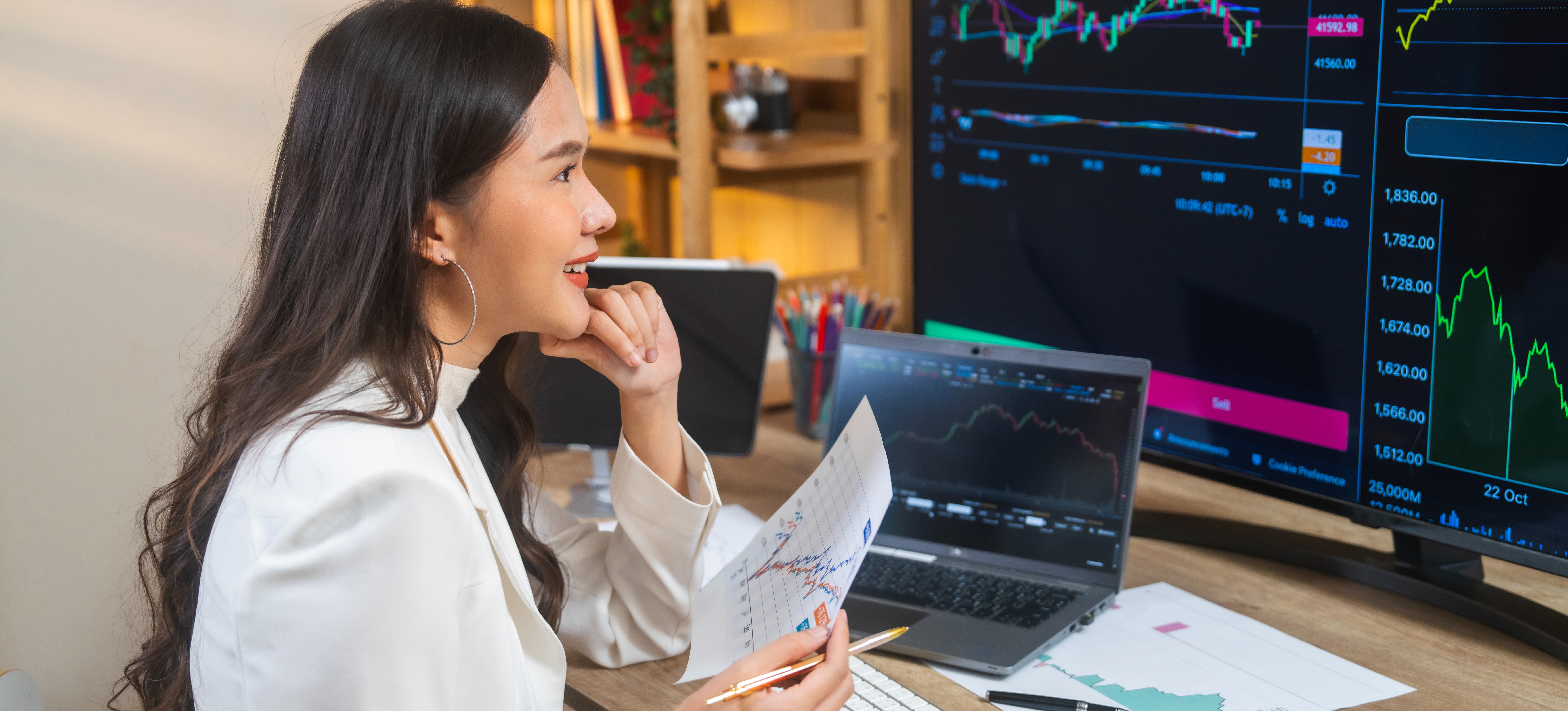 [Featured Image] A businessperson in an office setting smiles as they assess data integrity while looking at graphs displayed on their screen and documents.
