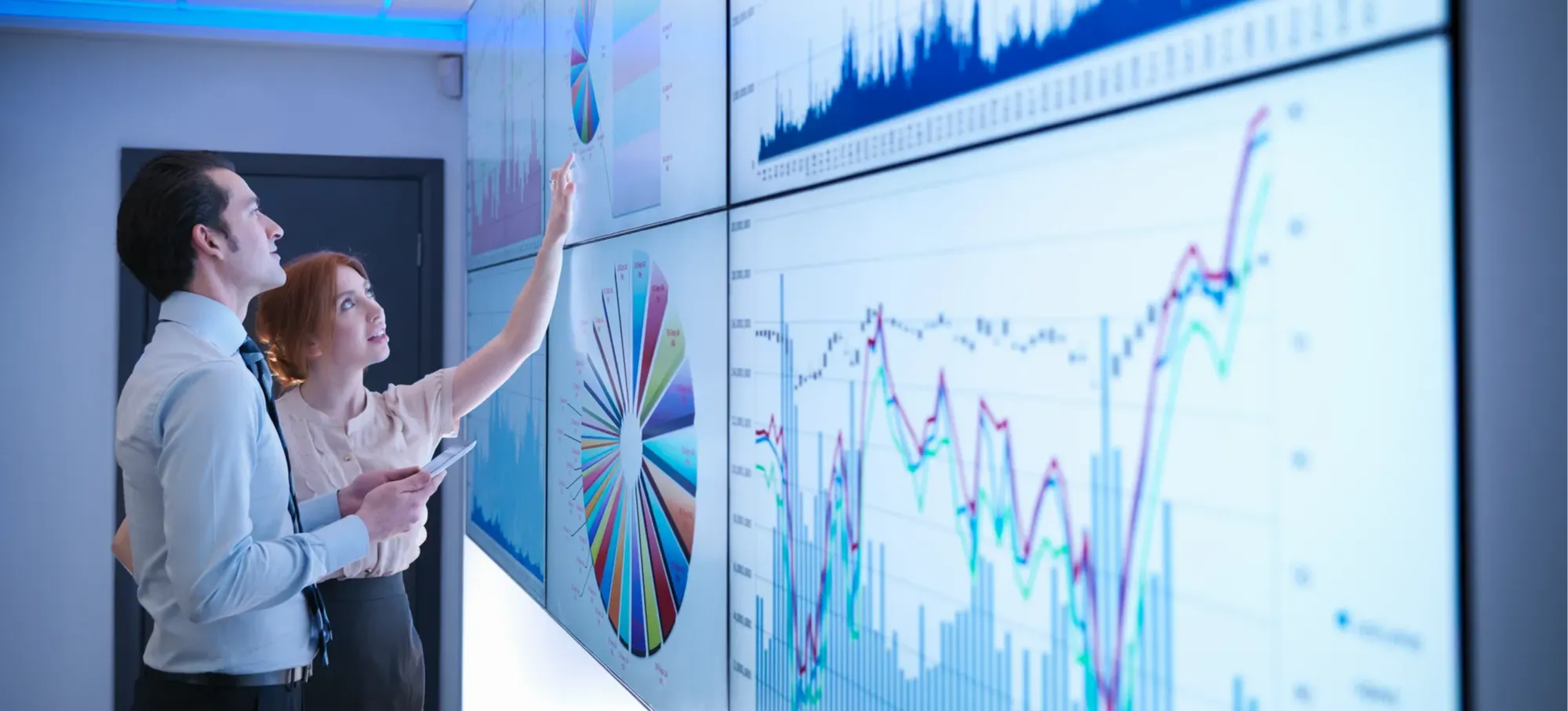 [Featured Image]: Two analysts review quantitative data displayed as charts and graphs across a collection of monitors.
