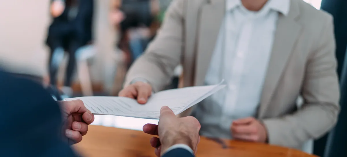 [Featured Image] An interviewee wearing a grey suit hands their email marketing resume to a potential employer in a job interview.
