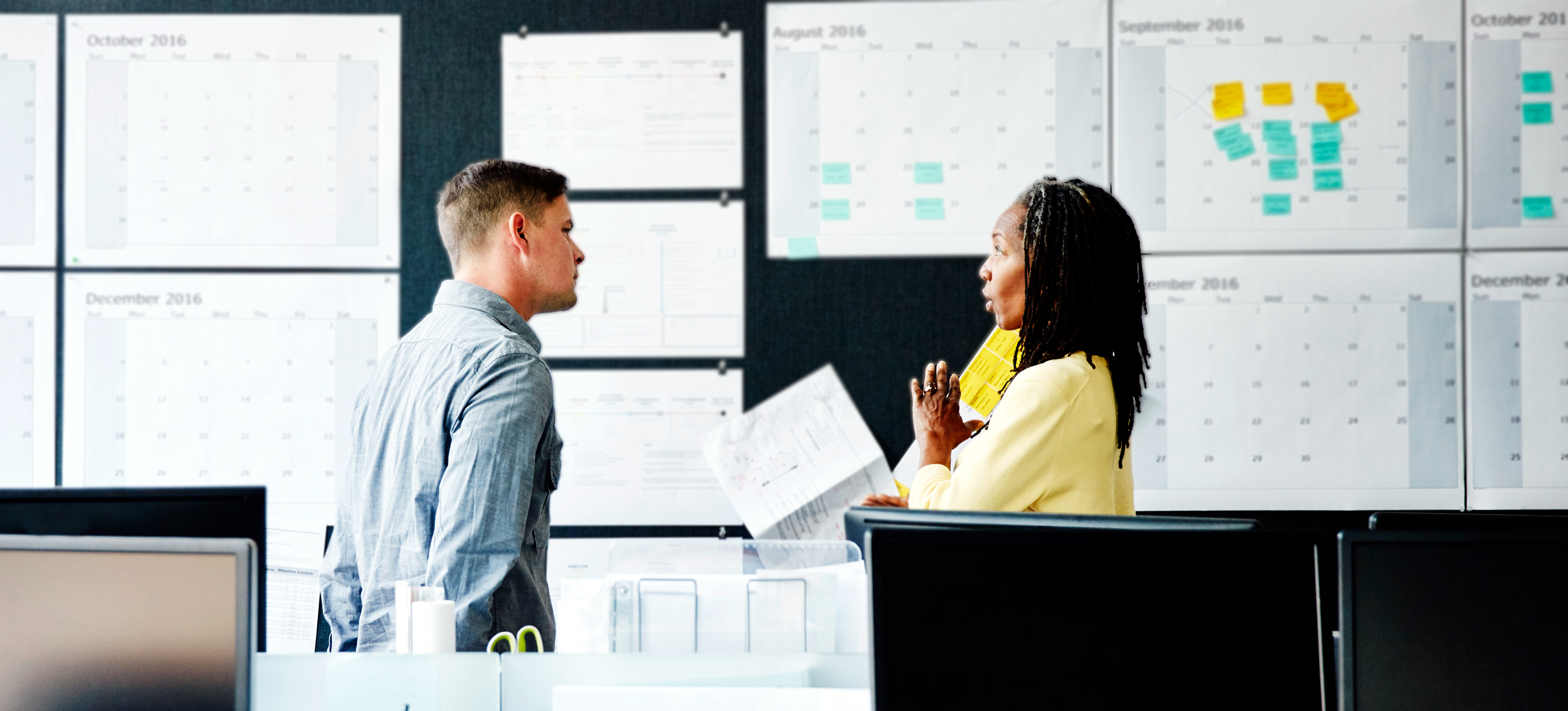 [Featured Image] An engineering manager in a yellow sweater is discussing the latest project with a design team member in a blue button-down. Both are standing in an open office, and large-format calendars cover the wall behind them.