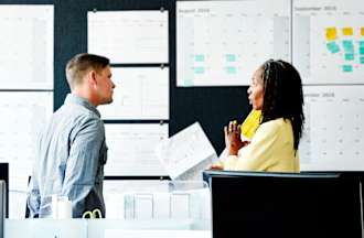 [Featured Image] An engineering manager in a yellow sweater discussing the latest project with a design team member in a blue button-down. They are standing in an open office and large-format calendars cover the wall behind them.