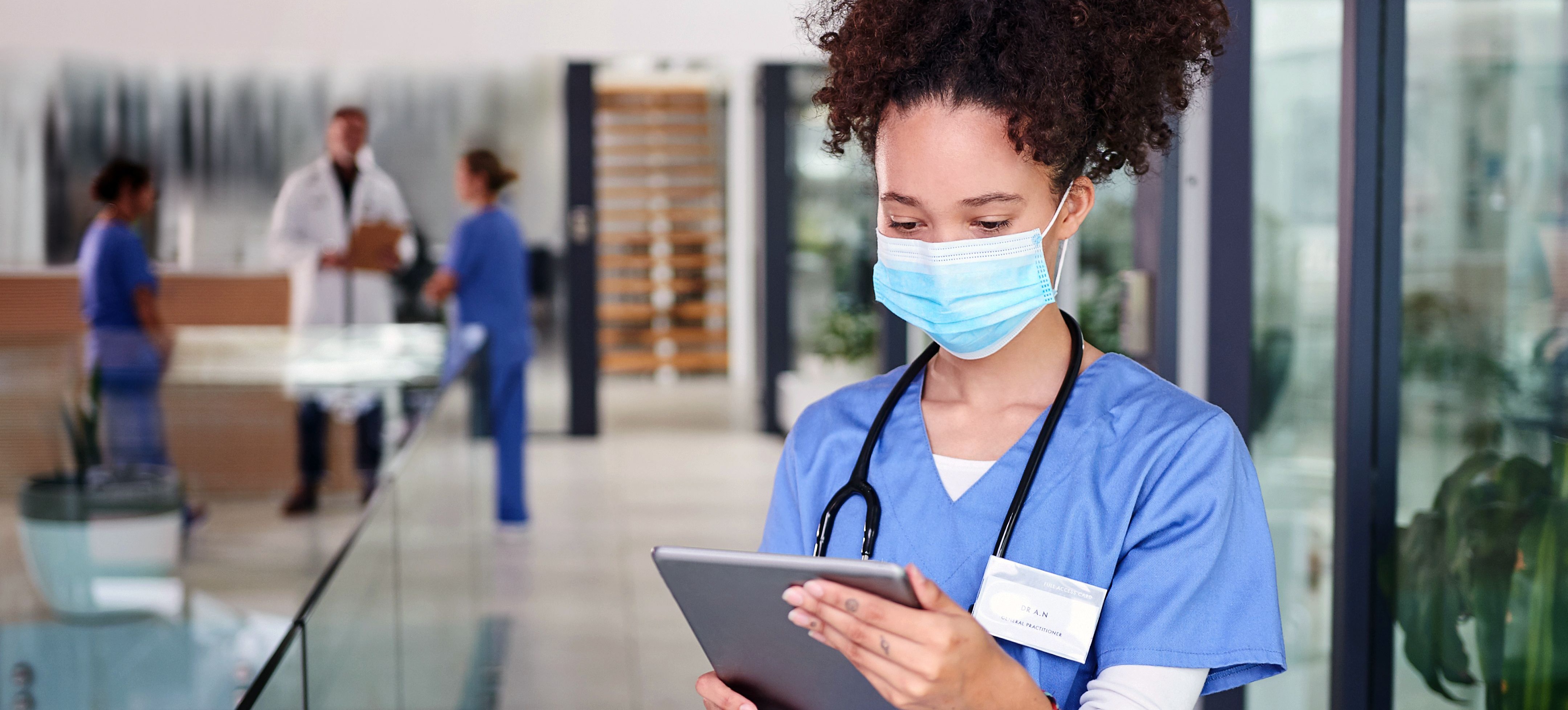 [Featured Image] A nurse wearing a mask and scrubs is using a tablet while standing in a hospital unit.