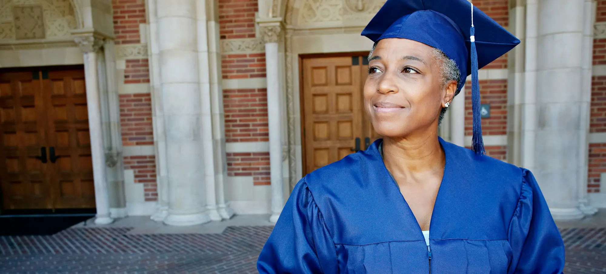 [Featured Image] A professional degree graduate in a blue cap and gown stands outside a university building after graduating with a business degree. 