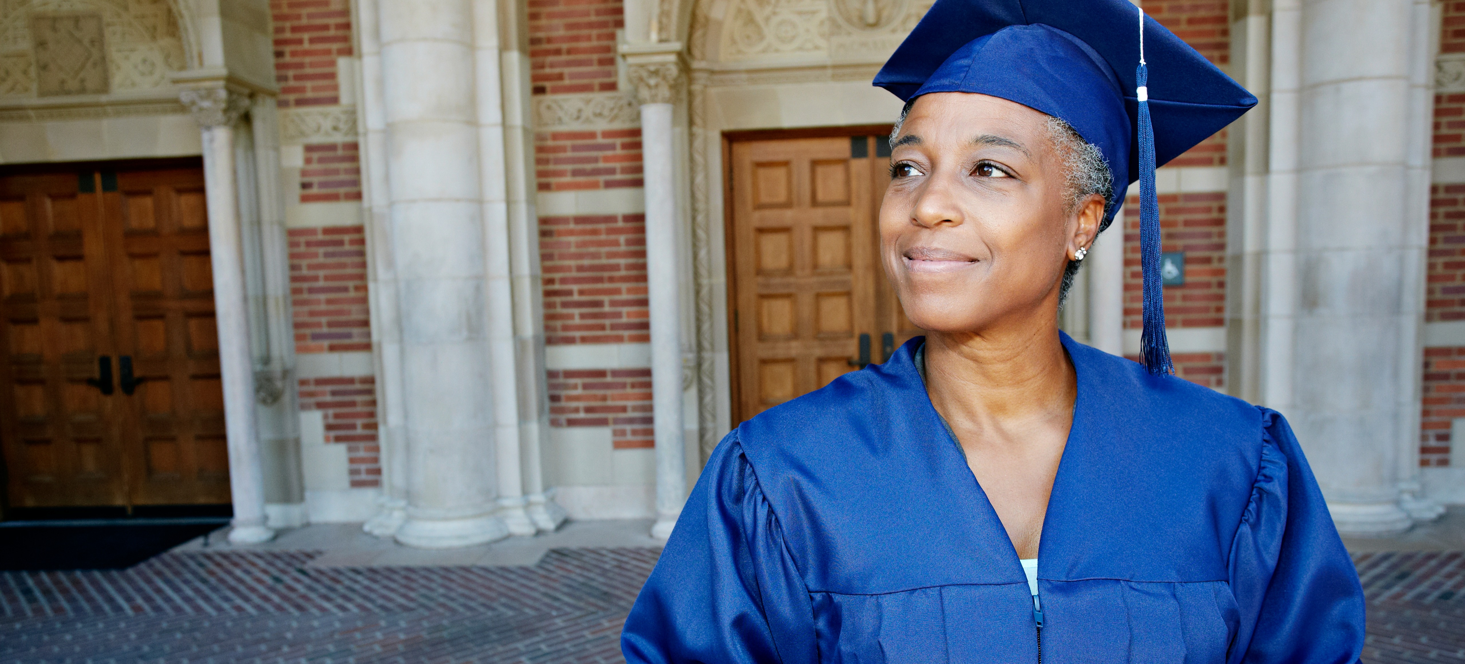 [Featured image] A professional degree graduate in a blue cap and gown stands outside a university building after graduating with a business degree. 