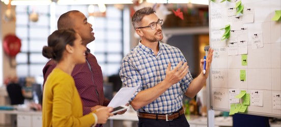 [Featured Image] Three coworkers discuss the contents of a whiteboard covered with sticky notes. 