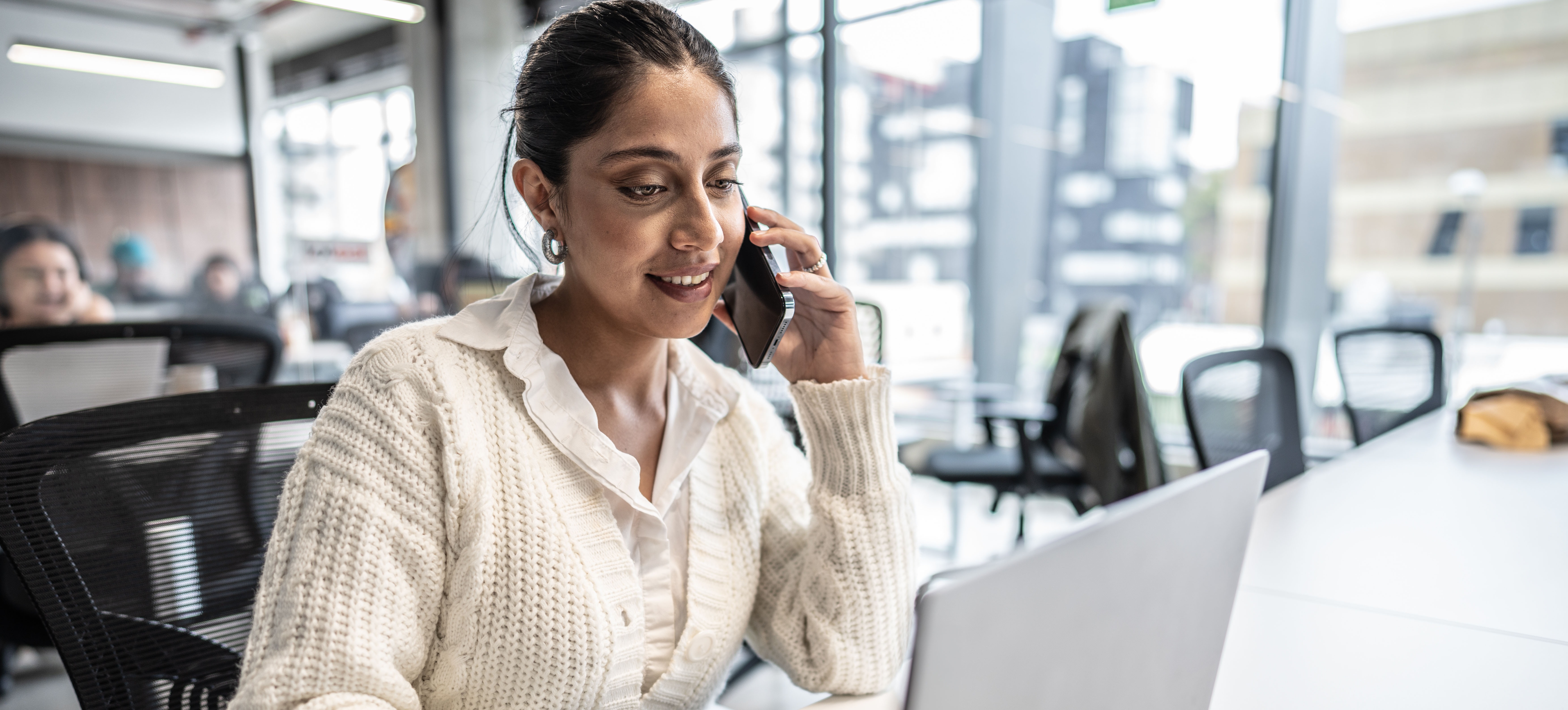 [Featured Image] A smiling coworker with an MA in communication talks on her smartphone while typing on a laptop in a busy office setting.
