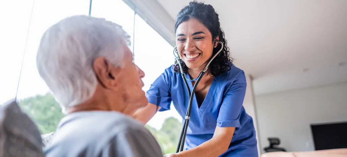 [Featured image] A smiling health care professional listens to their patient's heart through a stethoscope while they are in the patient's home.
