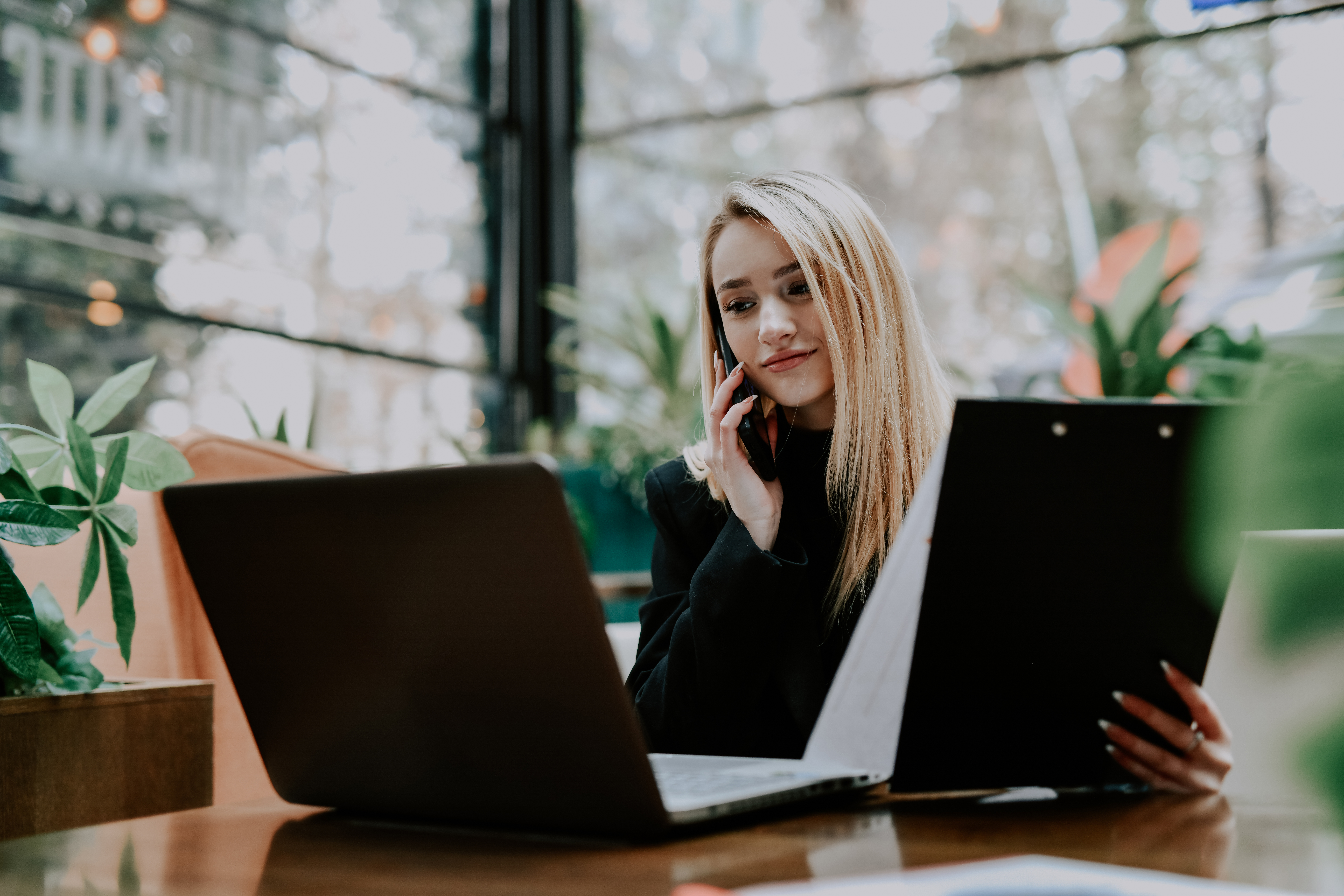 [Featured Image] An AI ethics professional talks on their smartphone while looking at their computer screen and some printed charts while working in a well-lit office with large windows behind them