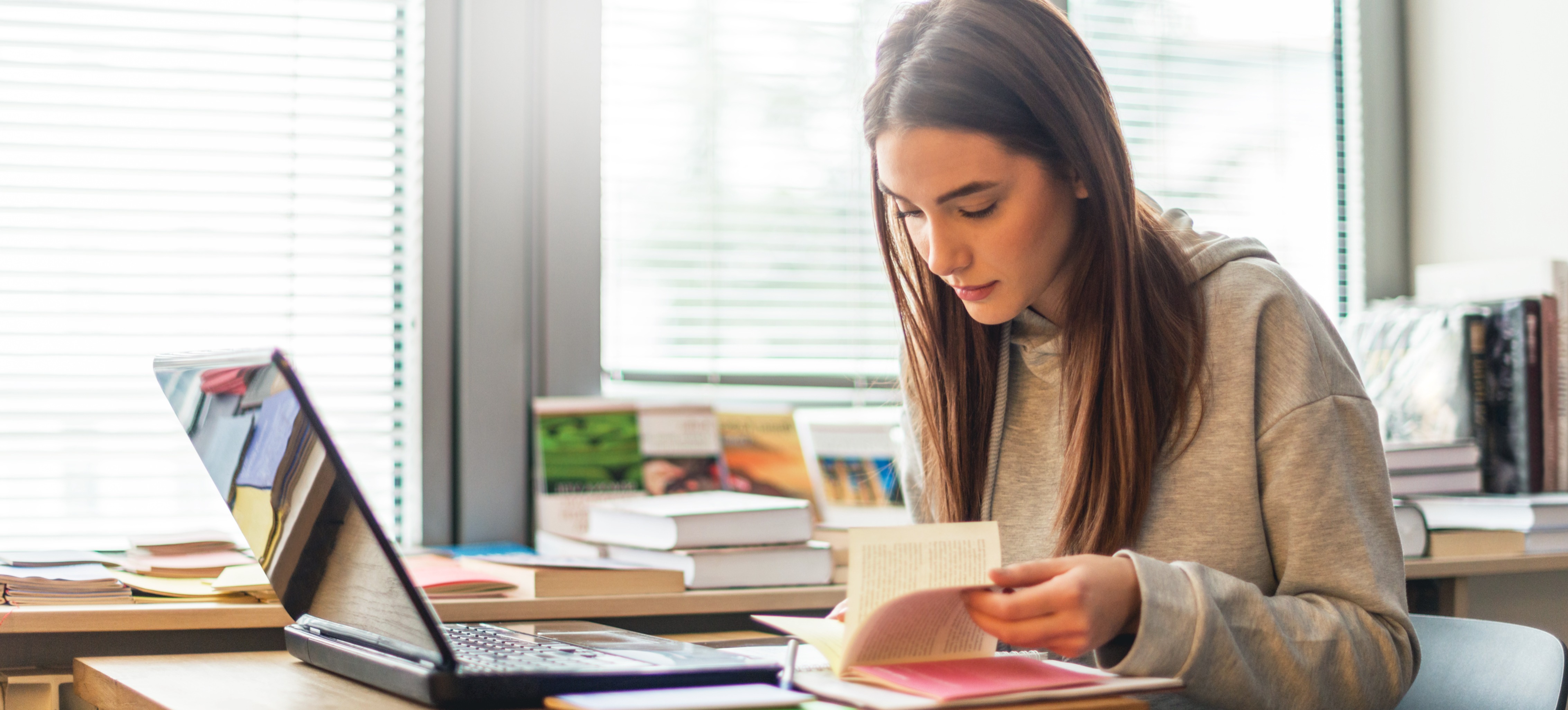 [Featured image] A woman wearing a gray hoodie reads a book in front of her laptop at her desk in front of sunlit windows.