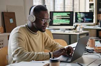 [Featured Image] An IT professional writes code at his desk in an office with several other workstations and computers. 