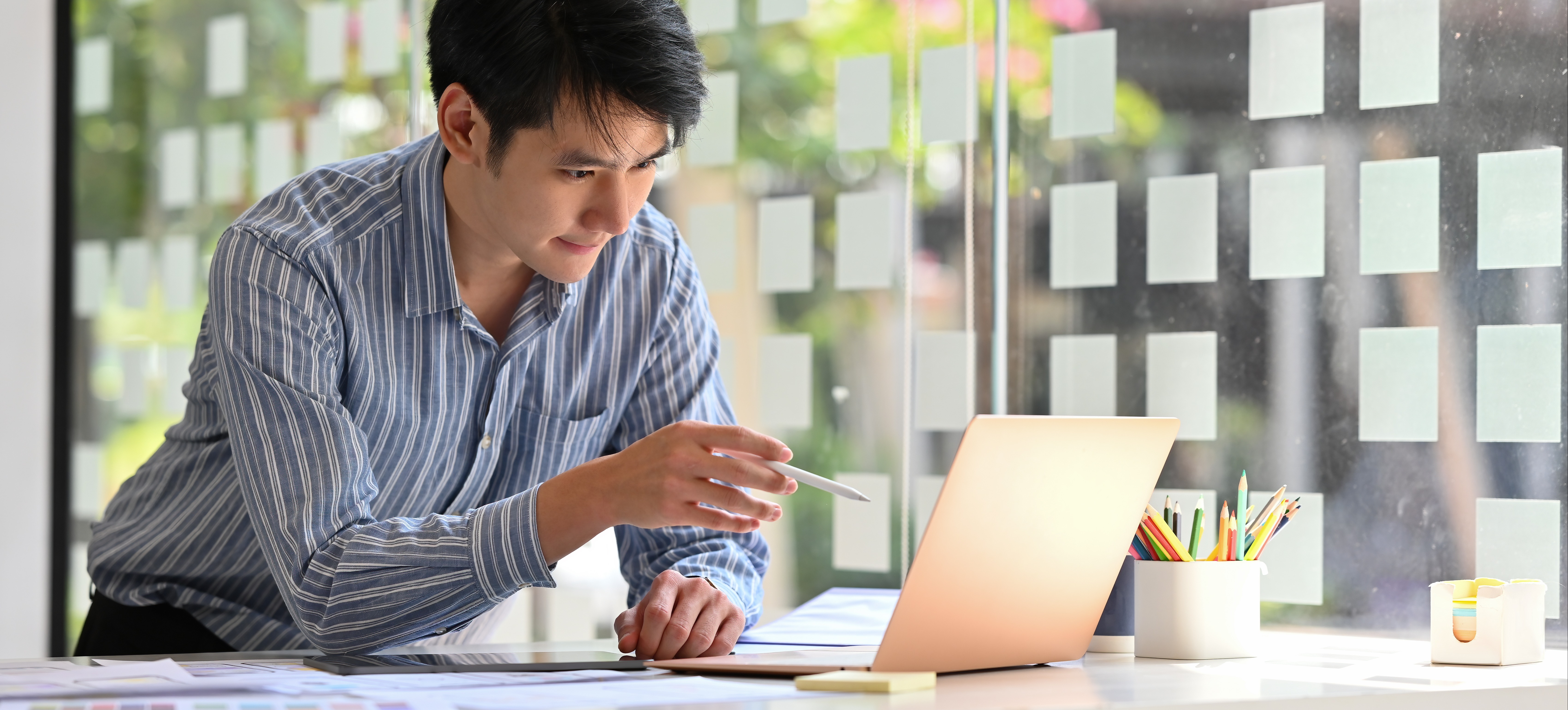 [Featured Image] An iOS developer in a blue collared shirt in the office at their standing desk using a stylus at a laptop as well as paper blueprints to design software applications for mobile phones.