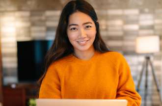 [Featured image] A young woman in a burnt-yellow sweater smiles into the camera while holding her laptop. 