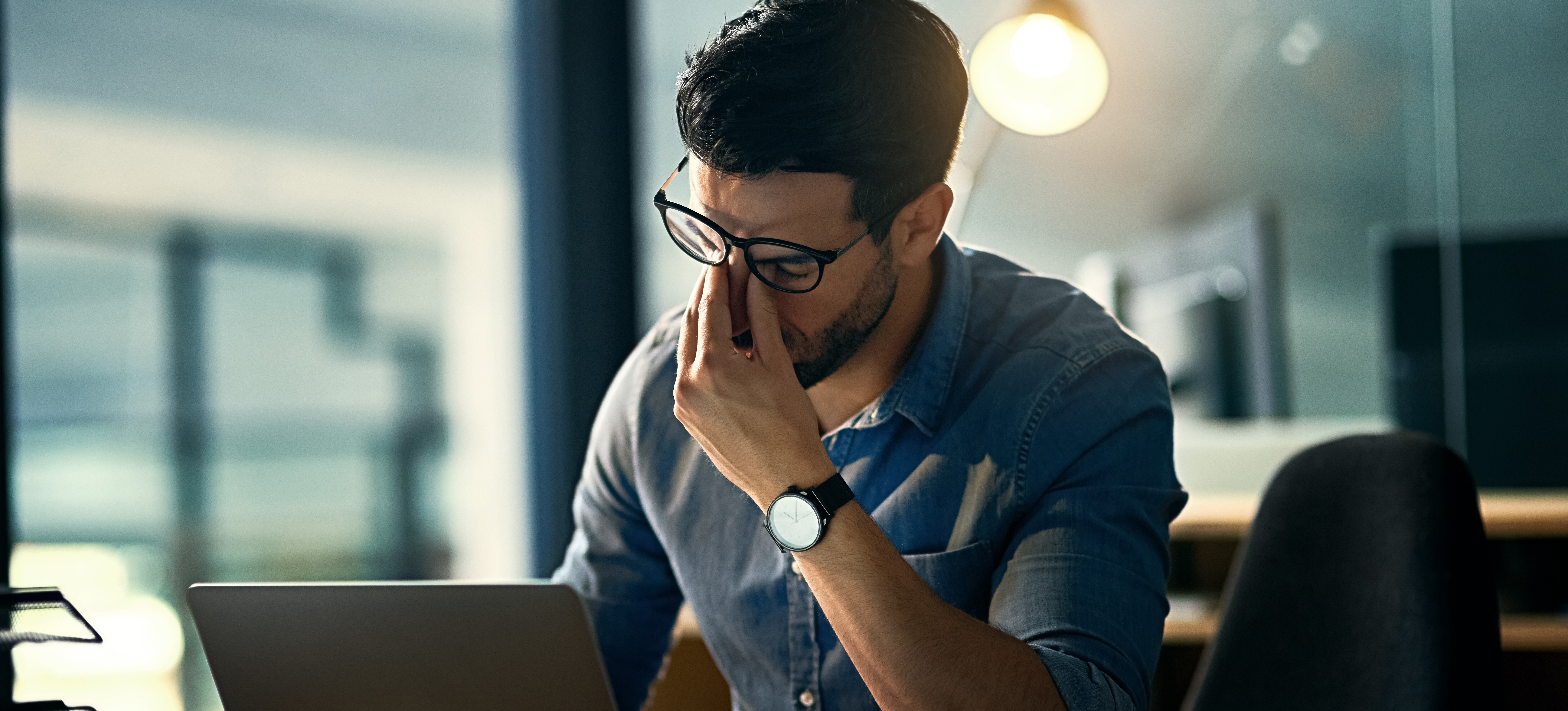 [Featured Image]: A person sitting in an office in front of a laptop rests their face in their hands as they experiences exhaustion at work.
