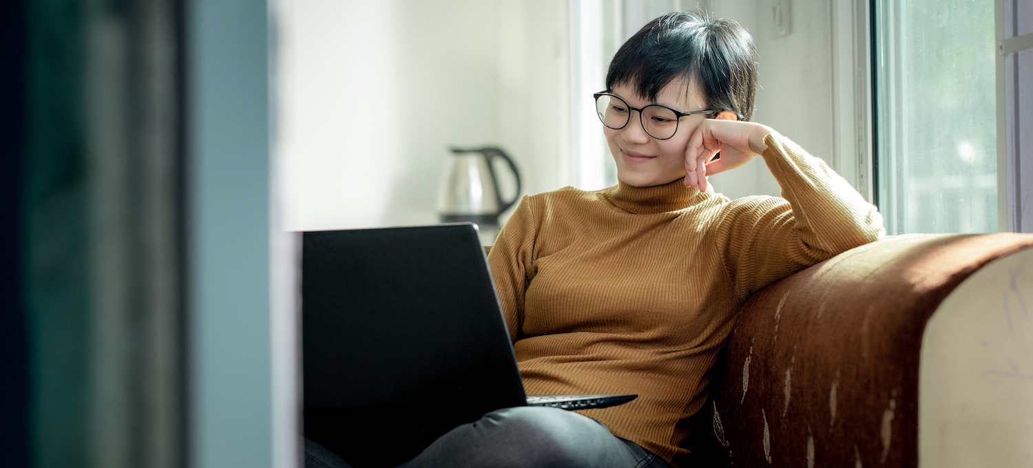 [Featured image] A young student with short hair and glasses sits on their couch smiling into their laptop.